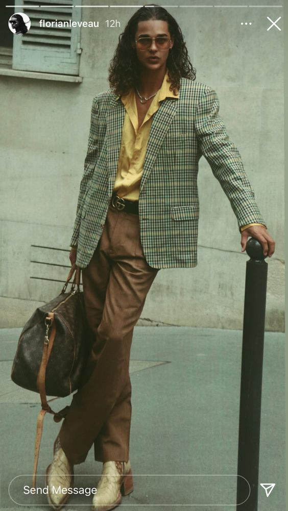 a stylish man in a plaid blazer, brown pants, and sunglasses leans against a wall in a city street while holding a leather bag. he is looking at the camera with a cool and confident expression.