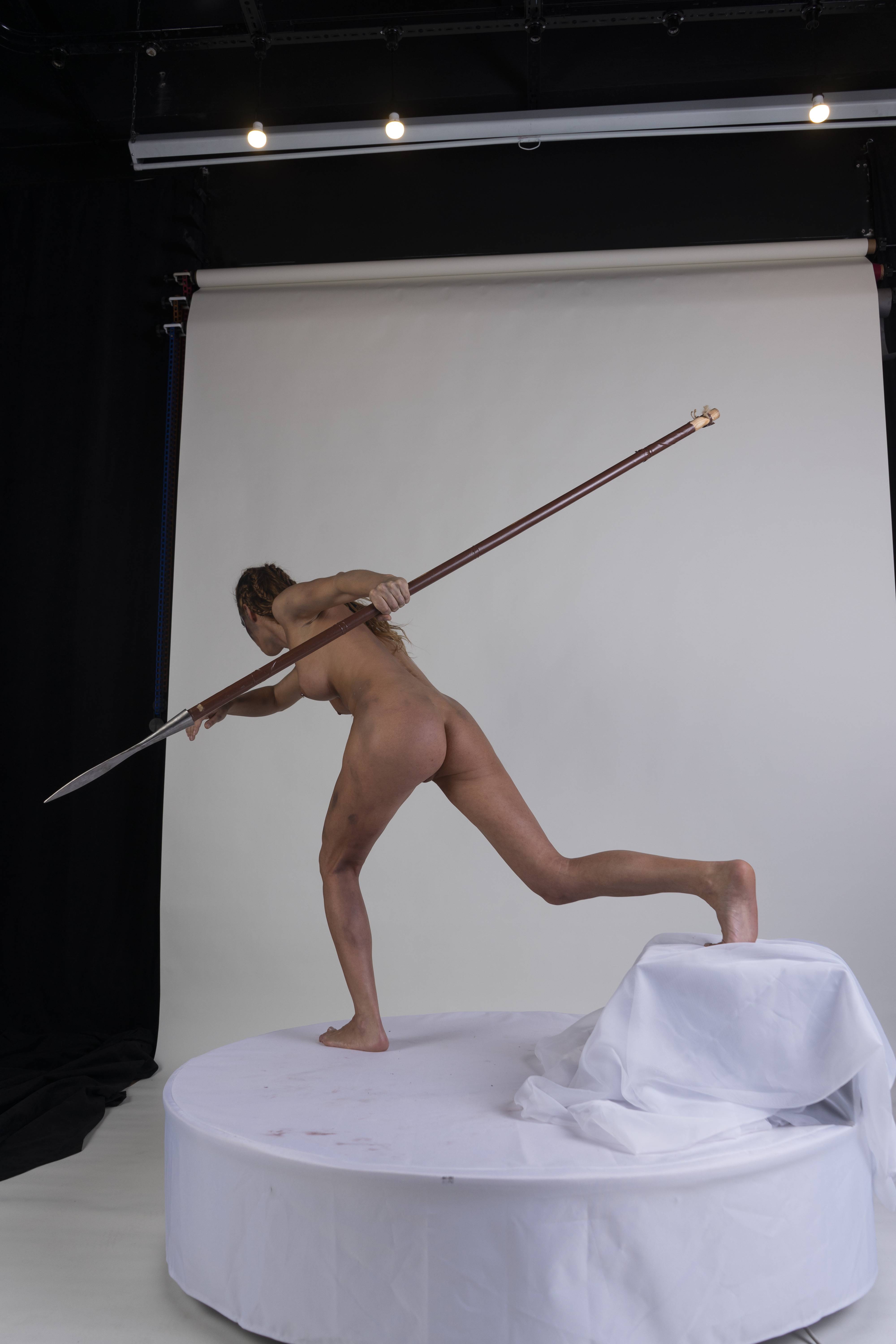 a nude woman stands on a white platform in a studio setting, holding a spear. she is barefoot, and her body is positioned in a dynamic pose as if aiming the spear. a white drape is visible on the platform. the background is a plain white screen.