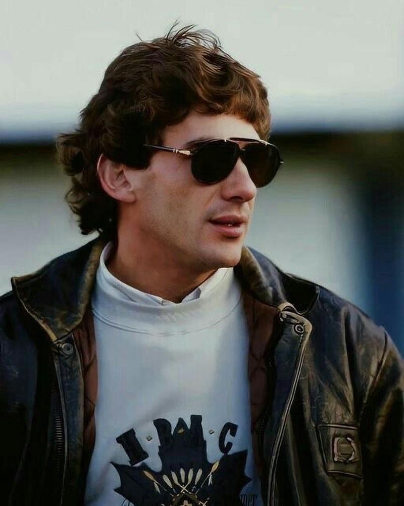 a man in sunglasses and a leather jacket looks away from the camera.  he is a racing driver.  the picture looks like a snapshot.