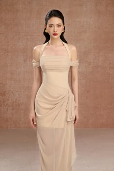 a beautiful woman in a beige halter neck evening dress is posing in a studio. the dress is fitted, with a long skirt. she has her hair pulled back, and is wearing earrings. she is looking directly at the camera. the background is a soft beige.