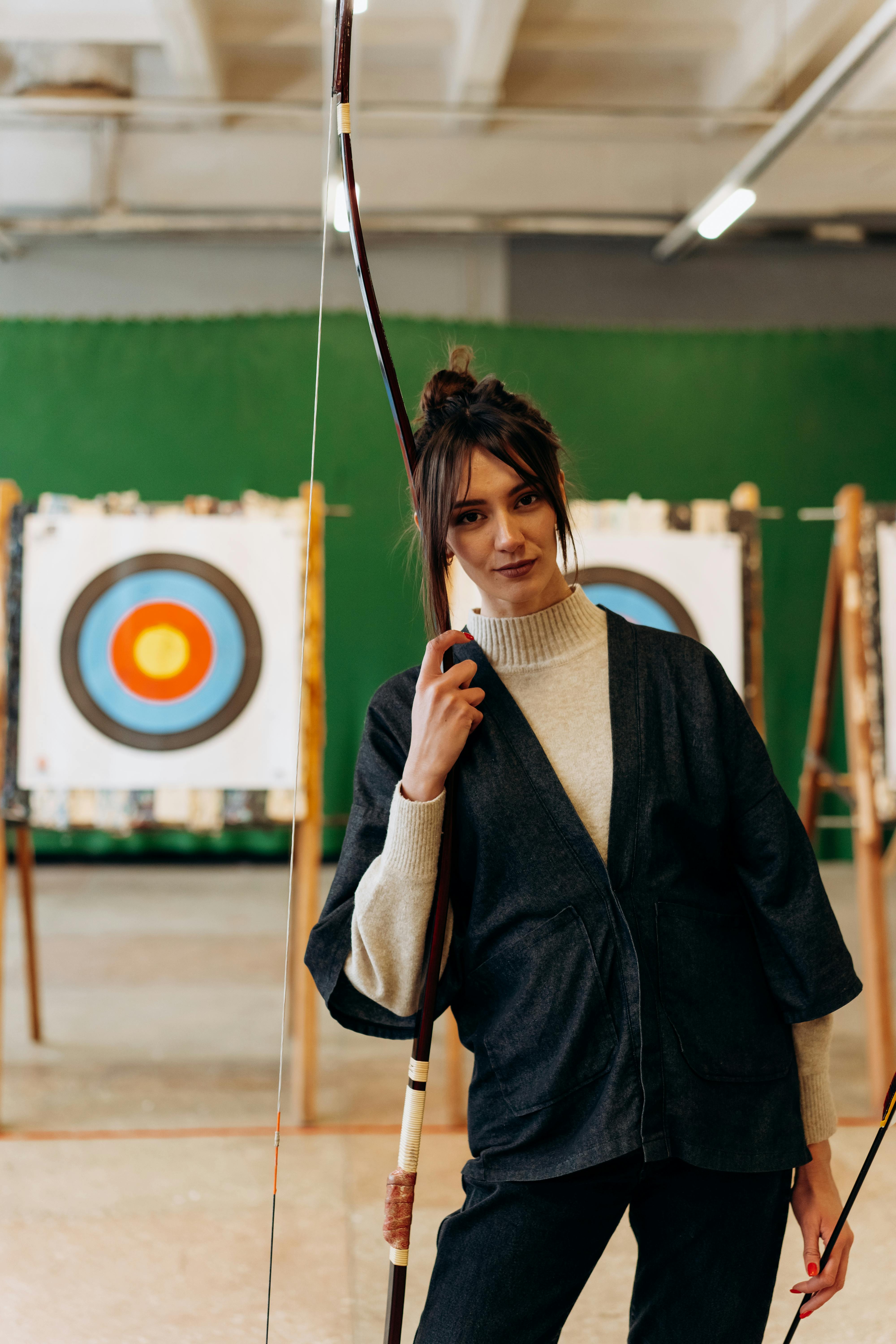 the image shows a young woman standing in an indoor archery range, holding a traditionalstyle japanese bow (likely a yumi used in kyudo). she's dressed in a stylish dark denim jacket over a creamcolored sweater and black pants, indicating a blend of modern and traditional attire.  she's positioned in front of a target, suggesting she's either preparing for or has just finished practicing archery. her focused expression conveys concentration and confidence. the overall ambiance is one of serenity and athleticism.