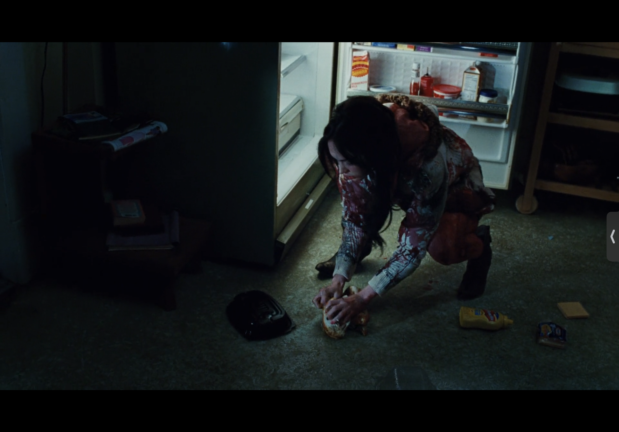 a woman in a bloody shirt is crouched on the floor of a dark kitchen, eating a sandwich next to an open refrigerator.  her face is obscured by her hair and the darkness of the room, but her bloody clothes and the overall messy state of the kitchen suggest that something disturbing has happened.  the image is evocative of a horror movie scene.