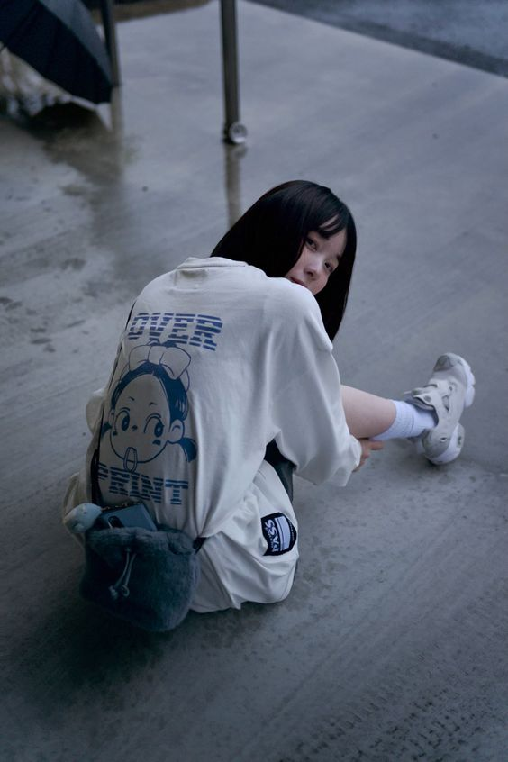 a young woman with dark hair is sitting on a wet sidewalk, posing for the camera. she is wearing an oversized white shirt with a graphic print, white socks, and white sneakers. she is also carrying a small gray plush bag. the background is a blurry urban scene with a wet sidewalk and overcast lighting.