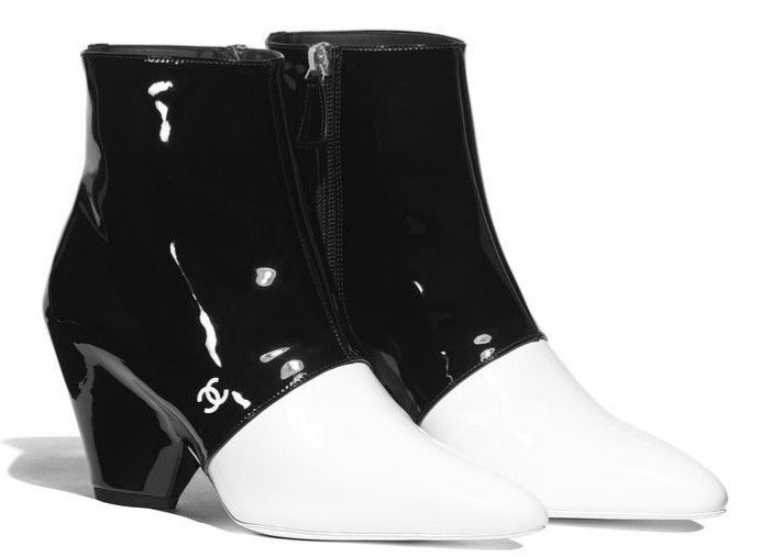 pair of chanel women's ankle boots are displayed. the boots are a twotone design, with the top half being black patent leather and the bottom half being white patent leather. the boots have a slight heel and a zipper closure on the inner side. the chanel logo is subtly embossed on the black leather portion of the boot. the background is white.