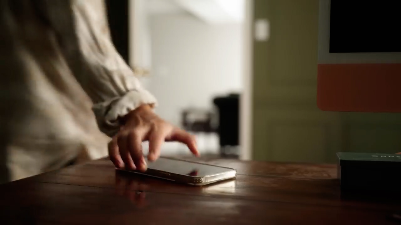 a closeup shot shows a woman's hand reaching towards a smartphone resting on a wooden desk. the background suggests a home office setting with warm, natural lighting. the overall ambiance is calm and inviting.