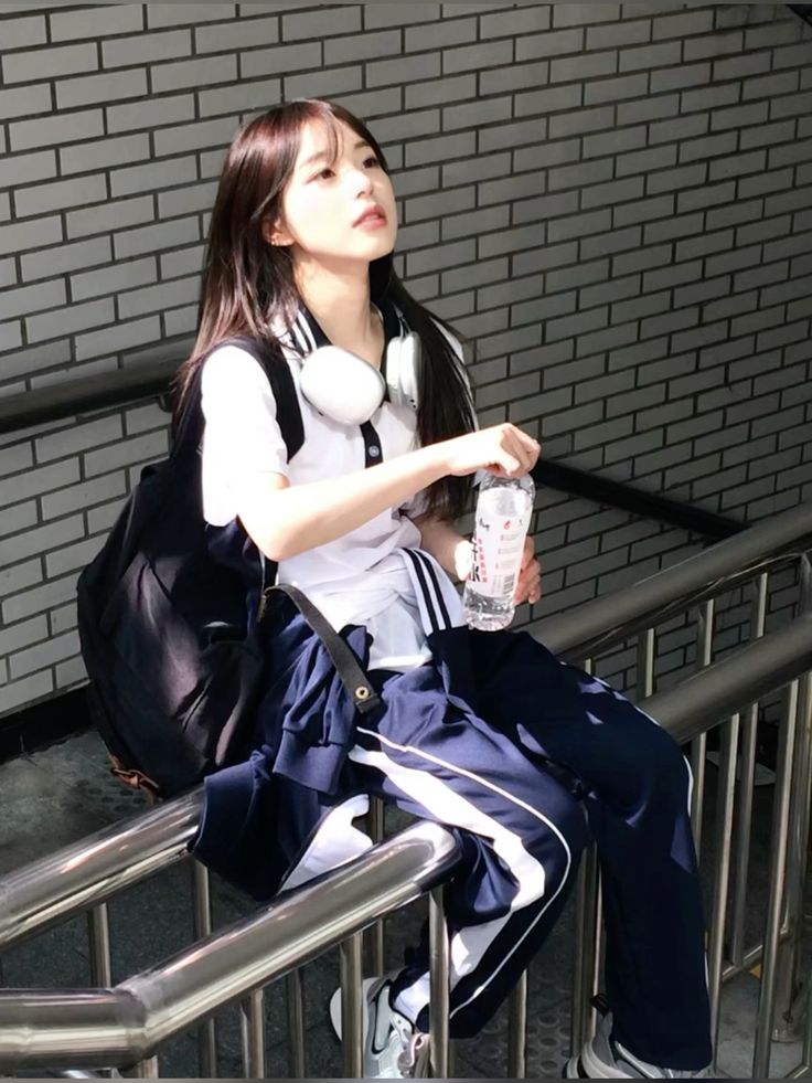 the image shows a young korean woman, possibly a student, sitting on a metal staircase outdoors.  she's wearing a classic school uniform consisting of a white polo shirt and navy blue track pants with white stripes down the sides.  she has a black backpack, white headphones around her neck, and is holding a bottle of water. the setting is a brick wall walkway, and sunlight illuminates her. her style is casual and fashionable.