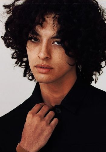 a closeup portrait of a young man with curly black hair. he is wearing a black shirt and looking directly at the camera with a serious expression. he has a strong jawline and piercing eyes. the lighting is soft and natural.