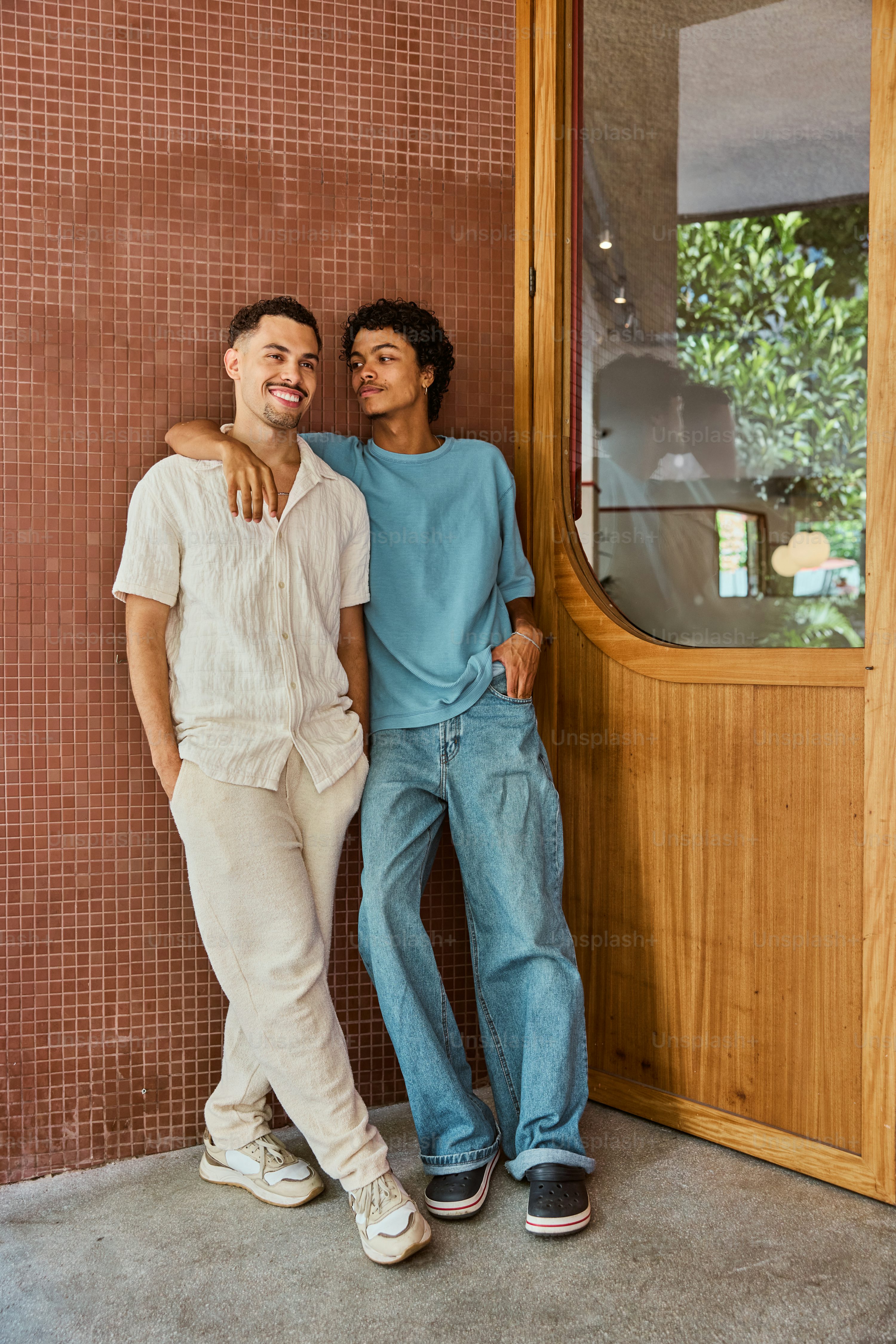 two young men stand close together, leaning against a redtiled wall beside a wooden doorway. one wears a white shirt and beige pants, the other a light blue shirt and jeans. they are smiling and appear relaxed and comfortable in each other's company. the setting appears to be an outdoor cafe or similar setting, with a large window visible in the background showing lush greenery. the overall mood is one of friendship, happiness, and togetherness.