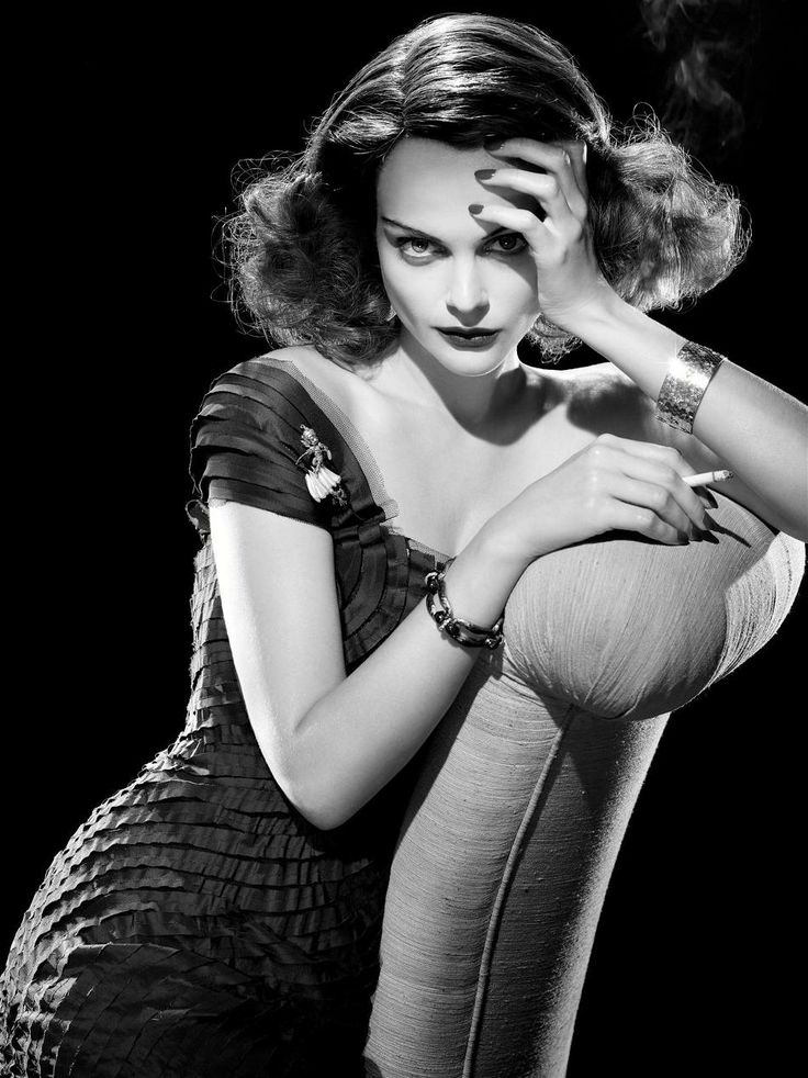 a black and white portrait of a glamorous woman holding a cigarette. she is wearing a dress, jewelry, and has her hand on her head.