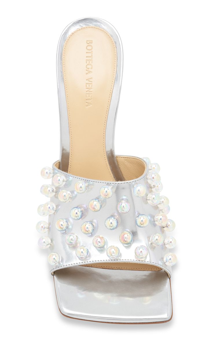 a closeup studio shot against a white background shows a pair of bottega veneta silver metallic iridescent pearl embellished slides. the shoes are squaretoed and feature numerous small, iridescent pearls adorning the upper. the insole is visible and displays the bottega veneta branding. the image showcases the detail and texture of the shoes, emphasizing their luxurious and glamorous aesthetic.