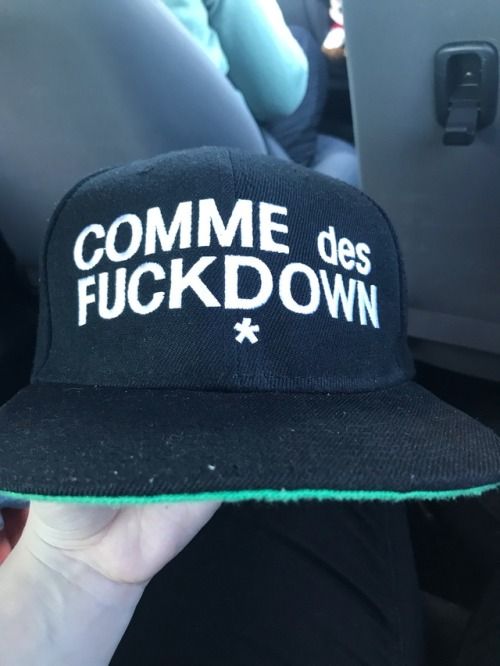 a closeup shot of a black snapback hat with white text "comme des fuckdown" and a small star symbol below. the underside of the brim is green. the hat is being held by a hand, and the background appears to be inside a car.