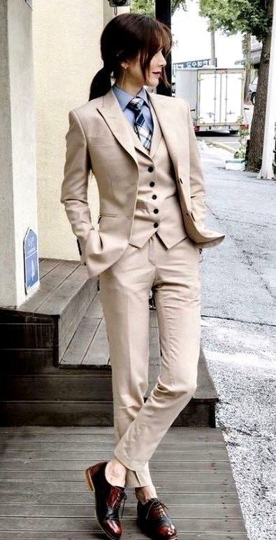 a fullbody shot of a stylish young asian woman wearing a light beige pantsuit.  the suit is a threepiece set, including a blazer, vest, and trousers. she's wearing a light blue and white patterned tie and dark burgundy and black oxford shoes. she has her brown hair pulled back into a ponytail. the setting appears to be an urban outdoor location, likely a city street.  the woman is standing and looking away from the camera, projecting a sophisticated and professional image.