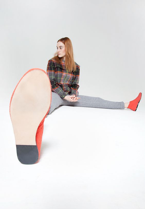 a young woman, dressed in a plaid shirt, grey pants, and red flats, is sitting on the floor with her legs outstretched. the photo is taken from a worm's eye perspective, focusing on the sole of her right shoe.