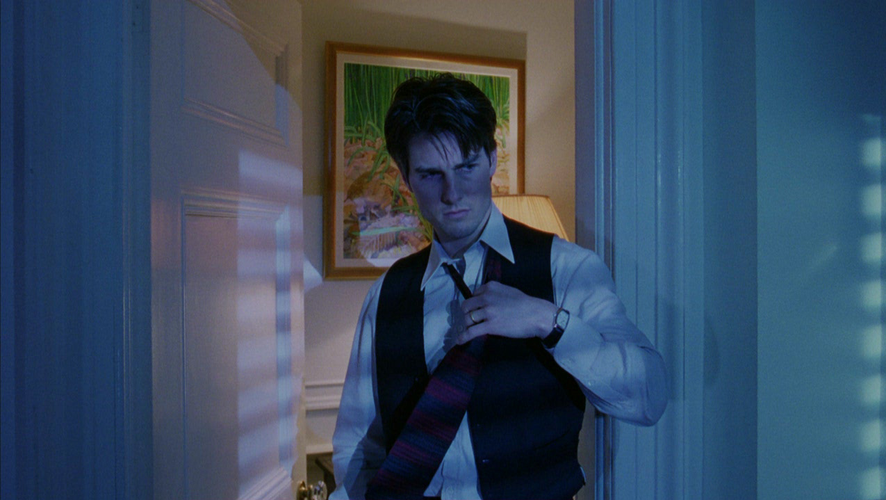 a young tom cruise, dressed in a white dress shirt, black vest, and patterned tie, stands in a dimly lit doorway. the blinds on the door cast vertical shadows across his face and torso, creating a sense of mystery and intrigue. the overall color palette is dark and moody, hinting at a tense or suspenseful scene.