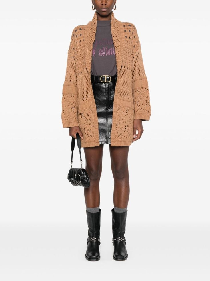 a woman wearing a brown knitted cardigan, a black leather skirt, black boots and carrying a black handbag. she's standing with her hands on her hips, in a studio.
