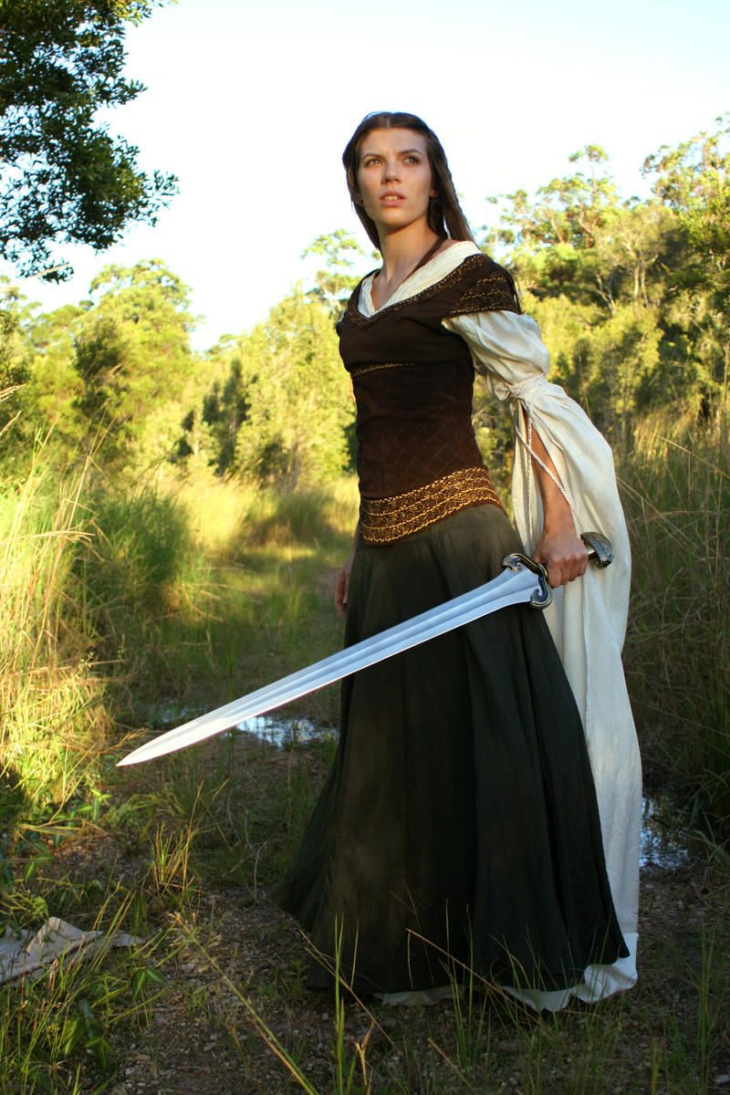 a fulllength shot of a woman dressed in a medievalstyle costume, holding a long sword. she's standing on a dirt path in a wooded area, with tall grass and trees surrounding her. the woman is looking away from the camera, and her expression is serious. the sunlight casts shadows in the background.  her dress is dark green and brown with offwhite/cream colored sleeves.