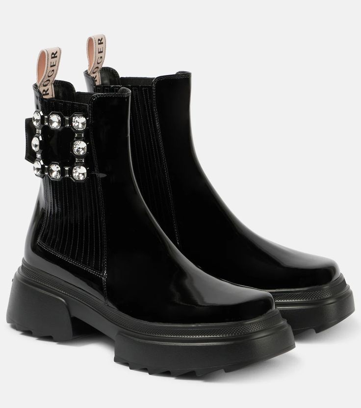 two shiny black ankle boots with lug soles are displayed against a white backdrop. the left boot has a prominent crystalencrusted buckle detail. both boots have beige pull tabs at the back with the brand name "roger" printed on them.