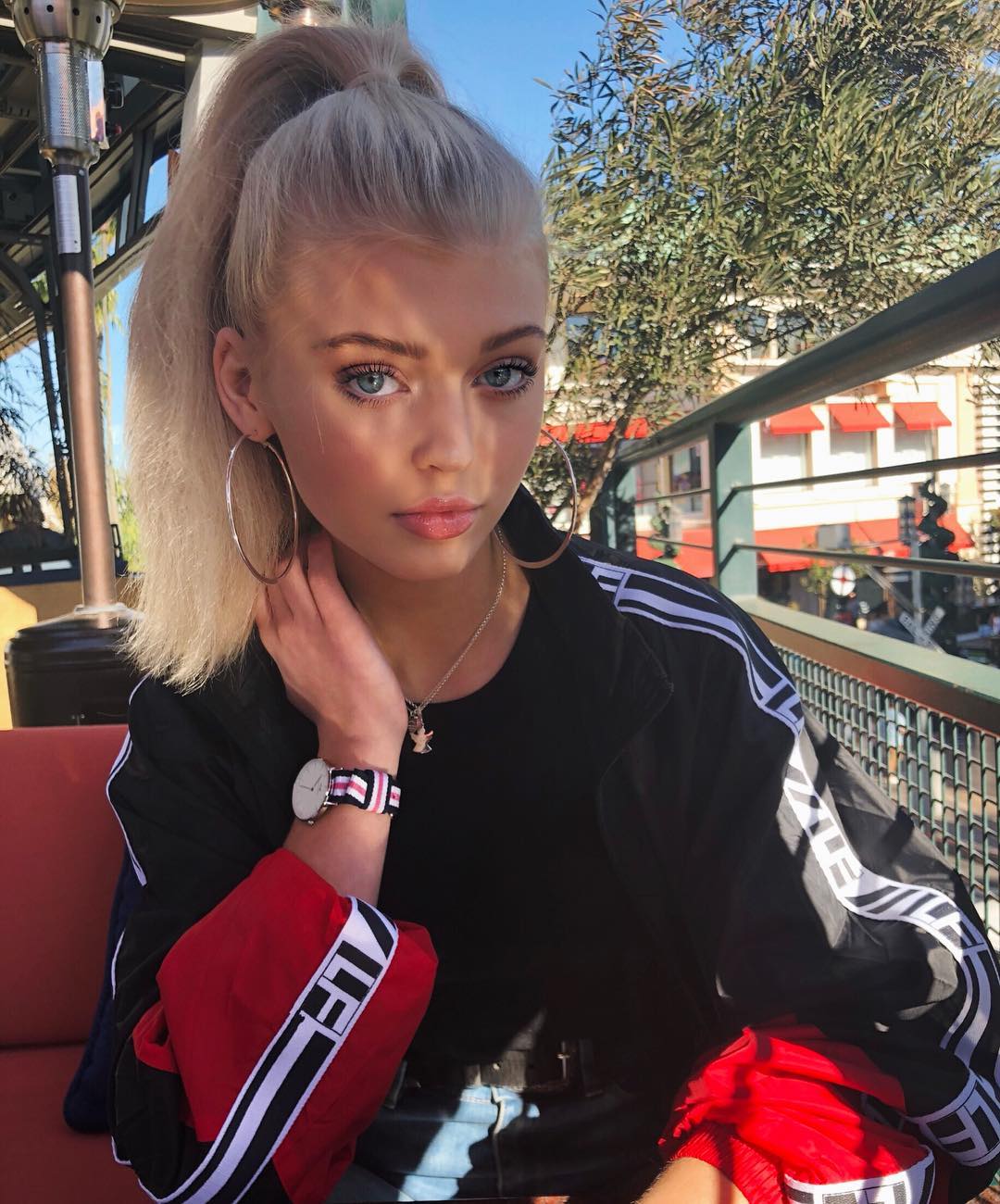a young woman with blonde hair pulled into a high ponytail, blue eyes, and wearing large hoop earrings, a necklace, and a watch is sitting outdoors. she is wearing a black jacket with red sleeves. in the background, there is a tree and a building.