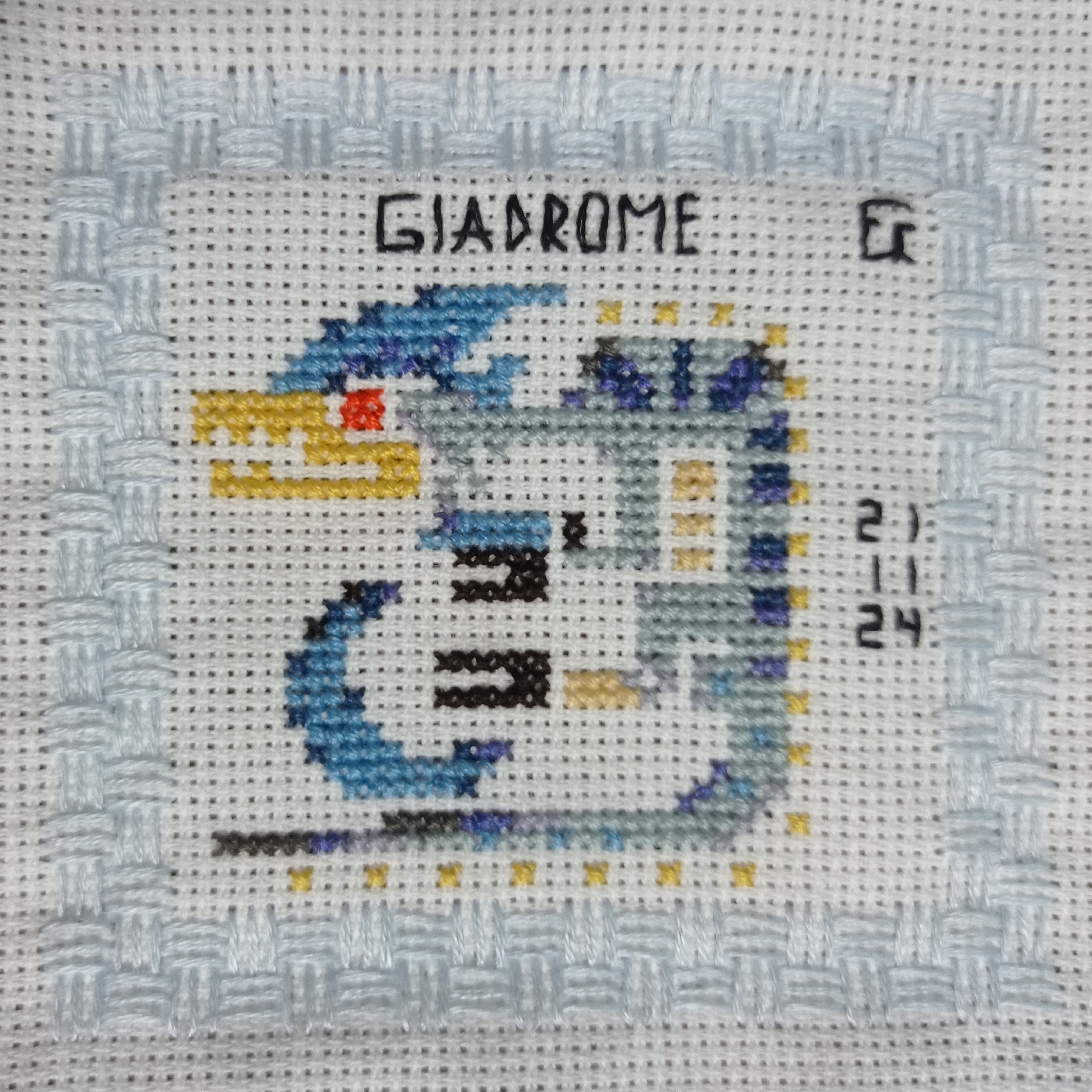 the image shows a closeup of a completed crossstitch piece. the design depicts a stylized creature, possibly inspired by the "giadrome" monster from the monster hunter video game series. the creature is rendered in shades of blue, gray, and yellow against a white background. the crossstitch work is neat and precise. the piece is bordered with a decorative lightblue woven stitch. the name "giadrome" is stitched into the top of the piece.  a small number sequence (2, 11, 24) is visible in the lower right corner of the design, which may be a project reference number or design code.