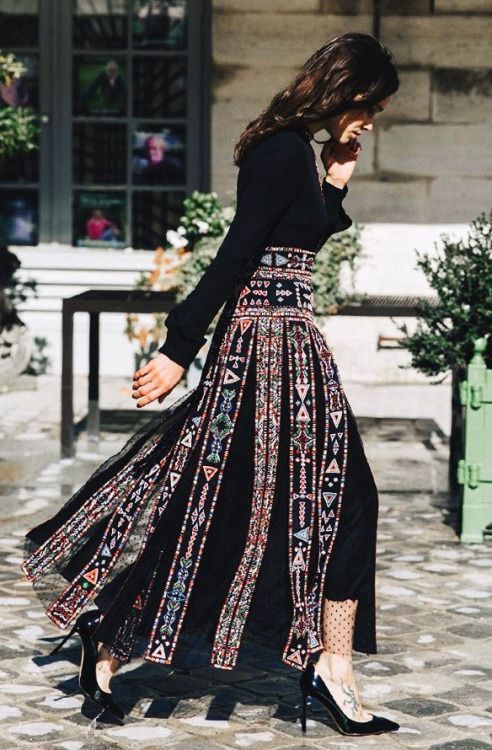 a woman is walking outside wearing a longsleeved black top and a long, black patterned skirt. she is also wearing black heels and has wavy hair. she has a tattoo on her foot. the background includes a walkway, a building, and some greenery.