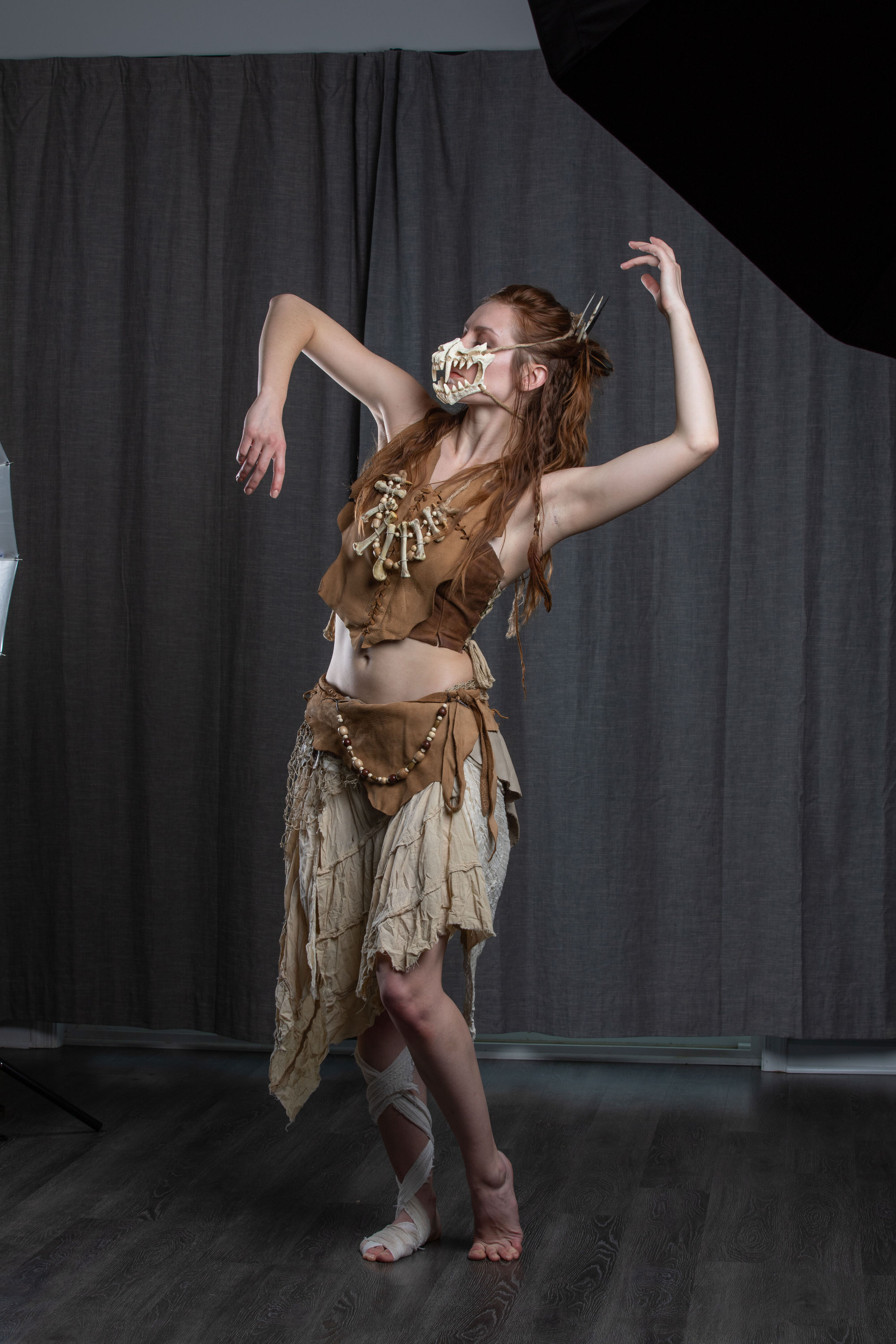 a female with long red hair pulled back in a halfup style dances in a studio setting. she is wearing a tribalinspired costume consisting of a beige wrap skirt, a brown top adorned with bones, and a bone mask covering her lower face. her right leg is wrapped in white fabric. the studio has a dark gray backdrop, and a softbox light source can be seen on the left side of the image. the overall ambiance suggests a creative portrait session with a focus on dance, costume, and artistic expression.