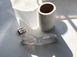 a pair of glasses sitting on a white table. there's also a glass cylinder and a roll of paper in the background. the whole scene is lit by natural light.