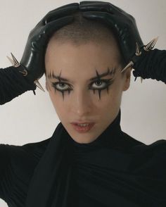 a portrait of a woman with a shaved head wearing black leather gloves with spikes and dark eyeliner. the gloves are on her head, and she is looking directly at the camera with a serious expression. she is dressed in black, and her hair is shaved off. her makeup is dramatic, with dark eyeliner and lipstick. she is sitting in a studio setting, and her background is a plain white wall.