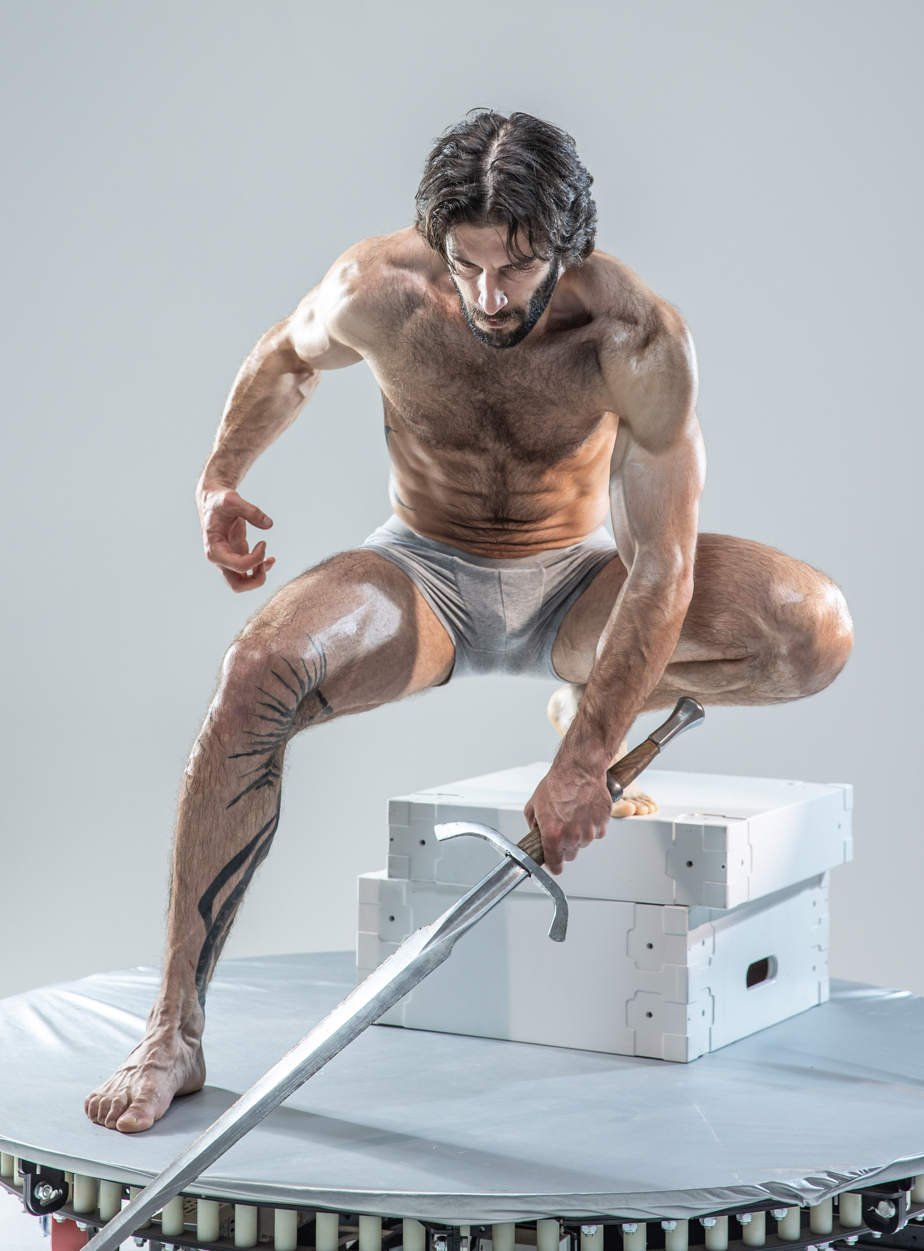 a muscular man with a beard and long hair is crouched on a white platform, holding a sword. he is shirtless and wearing gray underwear. the background is light gray. the man is illuminated by artificial light, creating a dramatic effect. the low angle perspective emphasizes his strength and power.
