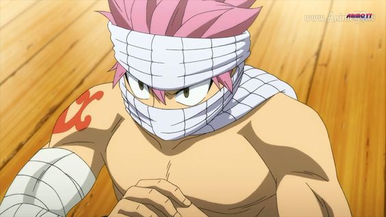 this closeup image showcases natsu dragneel, a prominent character from the anime fairy tail. he is shirtless, displaying a muscular build, with white bandages wrapped around his head, concealing his forehead and eyes, and on his left arm. his intense gaze and the red "x" mark on his right shoulder hint at a possible battle or challenge. the setting seems to be indoors, perhaps a training ground, illuminated by artificial lighting, which casts soft shadows and highlights his determined expression.