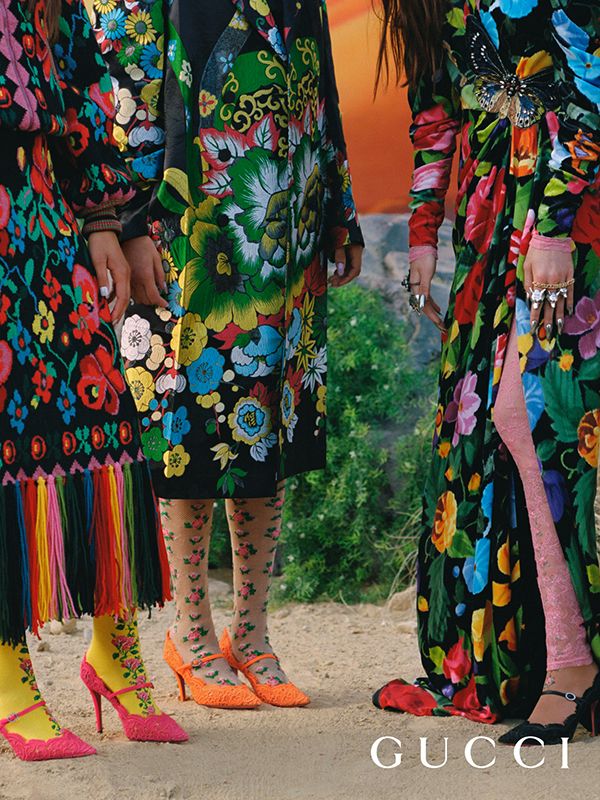 the image shows three models wearing vibrant, floral gucci clothing and accessories. they are standing outdoors on a dirt path with greenery in the background. each model wears a different floral garment, including a coat and a long dress. their shoes are orange and pink heels, and their tights have floral patterns. the overall style is eclectic and bohemian, with a focus on rich colors and intricate patterns. the gucci logo is displayed at the bottom of the image. the image suggests a fashion photoshoot for a spring/summer collection.