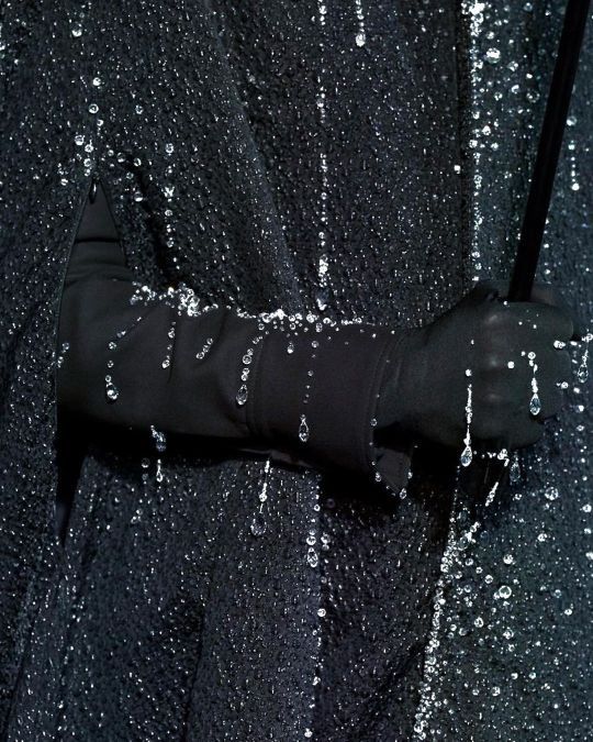 a closeup shot shows a blackgloved hand gripping an umbrella handle. the hand and a portion of the arm are visible against a backdrop of textured, dark fabric. water droplets cling to the glove, arm, and fabric, creating a glistening effect. the overall scene conveys a wet, possibly rainy atmosphere.