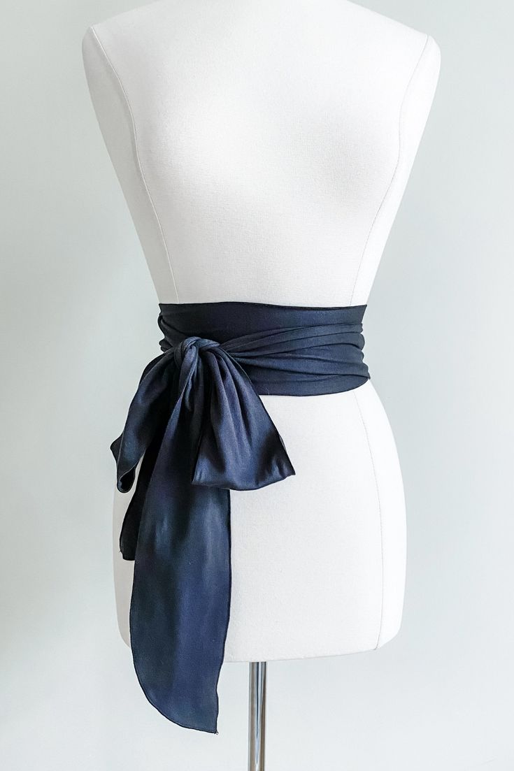 a navy blue silk sash is displayed on a white mannequin. the sash is tied in a bow at the front and is long enough to drape down the front of the dress.  the mannequin is a simple, white form against a clean white background. the sash is made of a lustrous, satinlike material. the overall impression is one of elegance and simplicity.