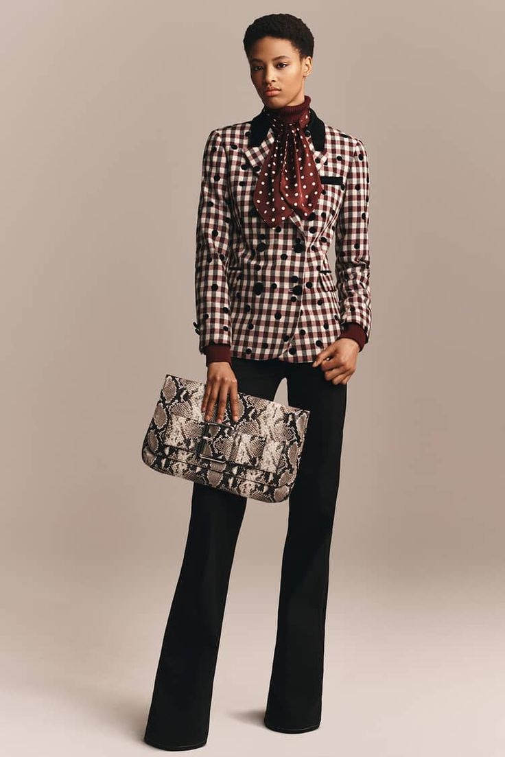 a woman wearing a checked black and red blazer with a red scarf and black pants is holding a snakeskin clutch and looking at the camera.