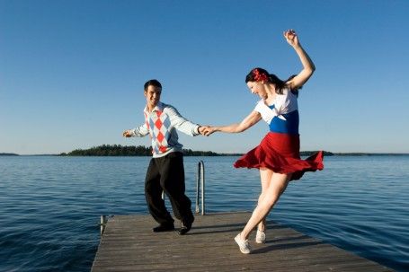 a couple dances on a dock overlooking a lake. the man is wearing a blue and white striped shirt and the woman is wearing a red, white and blue dress. they are both smiling and enjoying their time together.
