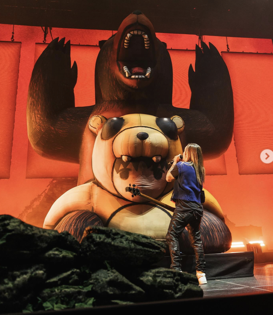 a musician dressed in black leather pants and a blue vest performs on stage in front of a gigantic inflatable bear and teddy bear. the bears are standing on the stage with their arms raised in a gesture of excitement. the backdrop is a red curtain. the performer is holding a bass guitar and singing into a microphone. the image is taken from a low angle, giving the viewer a good perspective of the stage and the inflatable bears.