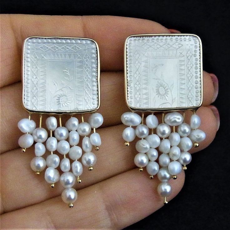 closeup photo of a pair of white pearl drop earrings. the earrings are made with a square white, engraved, carved top that is attached to a golden frame.  the square is connected to a golden chain, which is attached to many white round pearls.  the earrings are held in the palm of a hand.