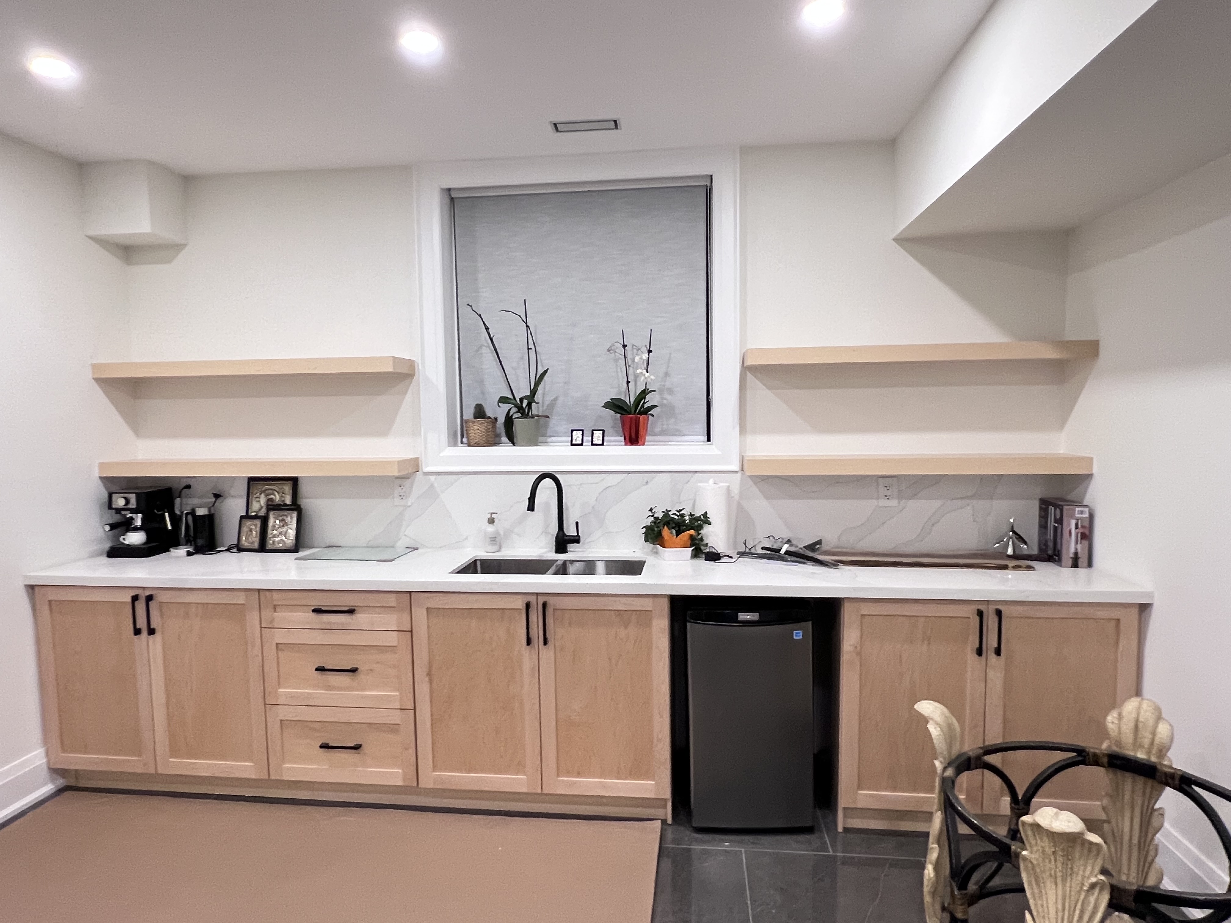 the image shows a newly remodeled or renovated small kitchen, likely in a basement.  the kitchen features lightcolored, possibly maple, wood cabinets with black hardware. a white countertop sits above the cabinets, which include a double sink with a black faucet and a small, integrated minirefrigerator.  three floating shelves, matching the cabinet color, are installed above the countertop. the walls are painted white, and recessed lighting provides illumination. a window with a lightcolored blind allows natural light into the space.  various small plants and kitchen appliances are visible on the countertops and shelves. the overall aesthetic is contemporary and minimalist.  a portion of a decorative chair is visible in the lower right corner. the flooring appears to be darkcolored tile.
