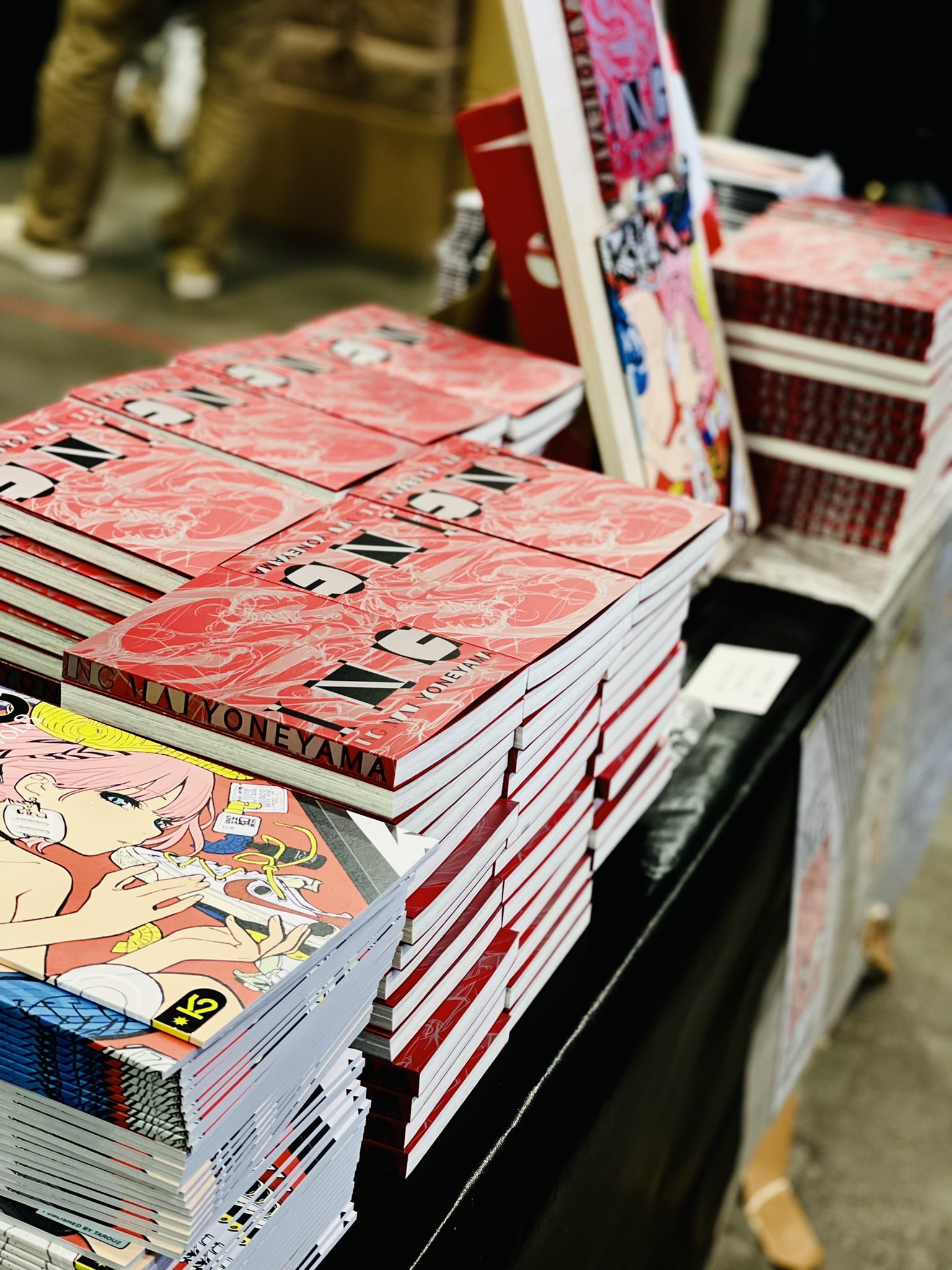 a closeup view of a table at an anime convention, showcasing stacks of independently published manga or art books. the books have vibrant red covers featuring japanese text and illustrations. the atmosphere is lively, with attendees browsing the merchandise and the artist's booth visible in the background. the lighting is bright and artificial, typical of an indoor convention hall.
