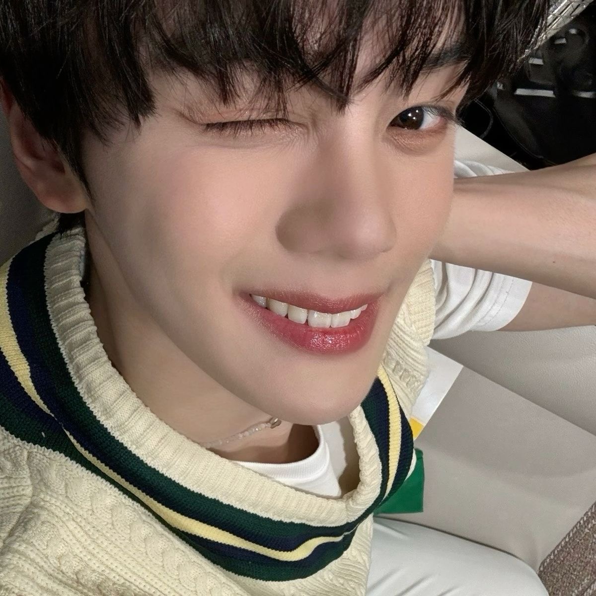 a closeup selfie of a young asian man with dark hair and brown eyes. he is wearing a white, green, and navy blue striped sweater and a silver necklace. he is smiling and winking at the camera. the lighting is soft and the perspective is from a high angle.  the background is blurred and suggests he is inside a car.