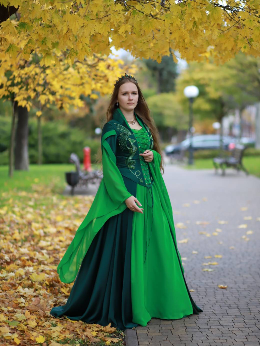 a woman in a medievalstyle green dress stands on a pathway in a park during autumn. yellow leaves cover the ground and hang from the trees overhead. she wears a tiara and jewelry, and her long hair flows down her shoulders. the dress has a detailed bodice and flowing sleeves, with a combination of light and dark green fabrics. benches and lampposts can be seen in the background.