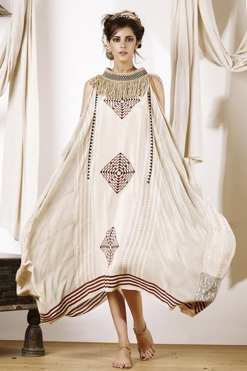 a woman stands in a studio setting, modeling a loosefitting, boho chic style dress. the dress is light tan in color and features ethnicinspired prints and patterns in dark brown and black. it has a tassel detail around the neckline and a fringelike effect on the shoulders. the dress also has a wide, flared shape and multiple horizontal stripes at the bottom edge. the woman is barefoot, wearing simple sandals, and jewelry on her head. draped fabric hangs in the background, contributing to the soft, bohemian vibe of the photo. a piece of dark wooden furniture sits to the left of the woman.
