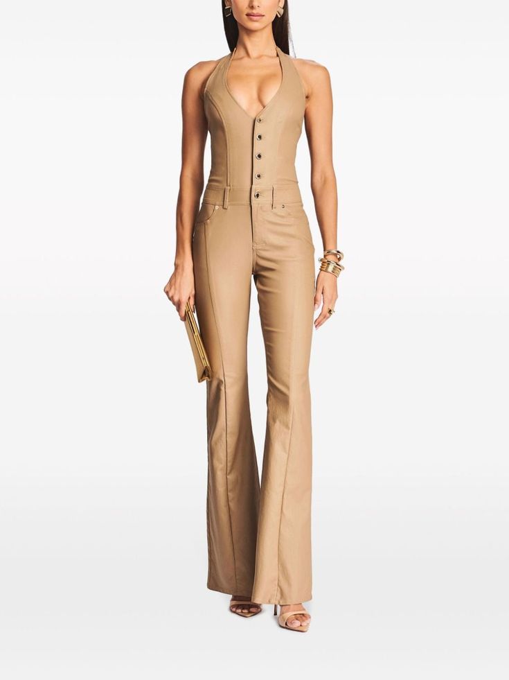 a woman model is standing in a studio against a white background. she is wearing a tan halter top jumpsuit with buttons up the front and flared legs. she is also wearing gold jewelry and heels. she is holding a gold clutch in her hand.