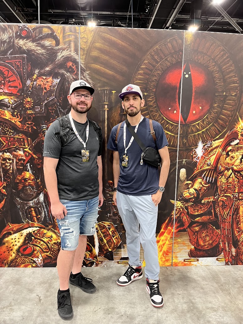 two men stand smiling in front of a large piece of warhammer 40k artwork at what appears to be a convention. the artwork depicts a space marine and an ominous eye. one man wears shorts and a tshirt, while the other wears lightcolored pants and a tshirt. both wear sneakers and hats. they appear to be enjoying the event.