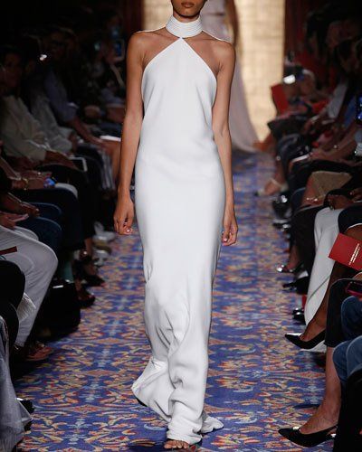 a model walks down the runway in a simple yet elegant white halter neck gown. the dress is fitted and flares out slightly at the bottom, creating a graceful silhouette. the model is walking confidently, showcasing the dress's beauty. she is surrounded by an audience of people who are watching the fashion show. the runway is adorned with a patterned carpet.