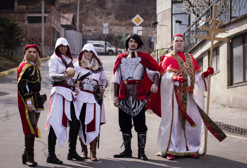a group of people are standing on a road, dressed in elaborate costumes reminiscent of characters from the video game "assassin's creed." they include a character in white robes with a hood, a character in renaissance clothing with a red beret, a character with a red cape and silver armor, and a character dressed as a pope with a cross. the setting appears to be a street with buildings and vehicles in the background.