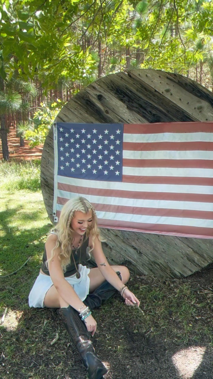 a blond woman is sitting on the grass in front of an american flag attached to a wooden cable reel in an outdoor setting. she is wearing an olive green tank top, denim shorts, and black boots. she is also wearing jewelry including bracelets and a necklace. the background includes woods, trees, and grass.