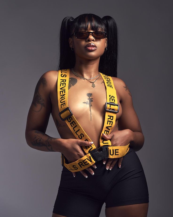a young woman wearing black shorts and yellow suspenders with "sells revenue" written on them. she's also wearing sunglasses and has her hair in pigtails.  the woman is posing confidently for the camera in a studio setting with a plain grey background.