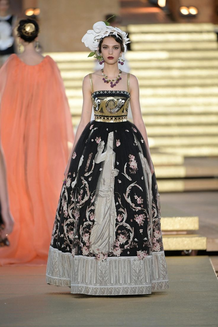 a model walks down a runway in a black gown with gold embroidery and a white floral headpiece. the dress is adorned with delicate floral embroidery, and the model is accessorizing with gold jewellery. the gown is a statement piece of haute couture fashion, and the model's confident pose and graceful walk make her a real headturner.
