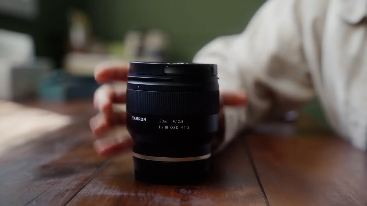 a tamron 20mm f2.8 lens is placed on a wooden table and a person's hand is reaching for it. the lens is in focus and the background is blurred, creating a shallow depth of field. the lighting is soft and natural, coming from the left side of the image.