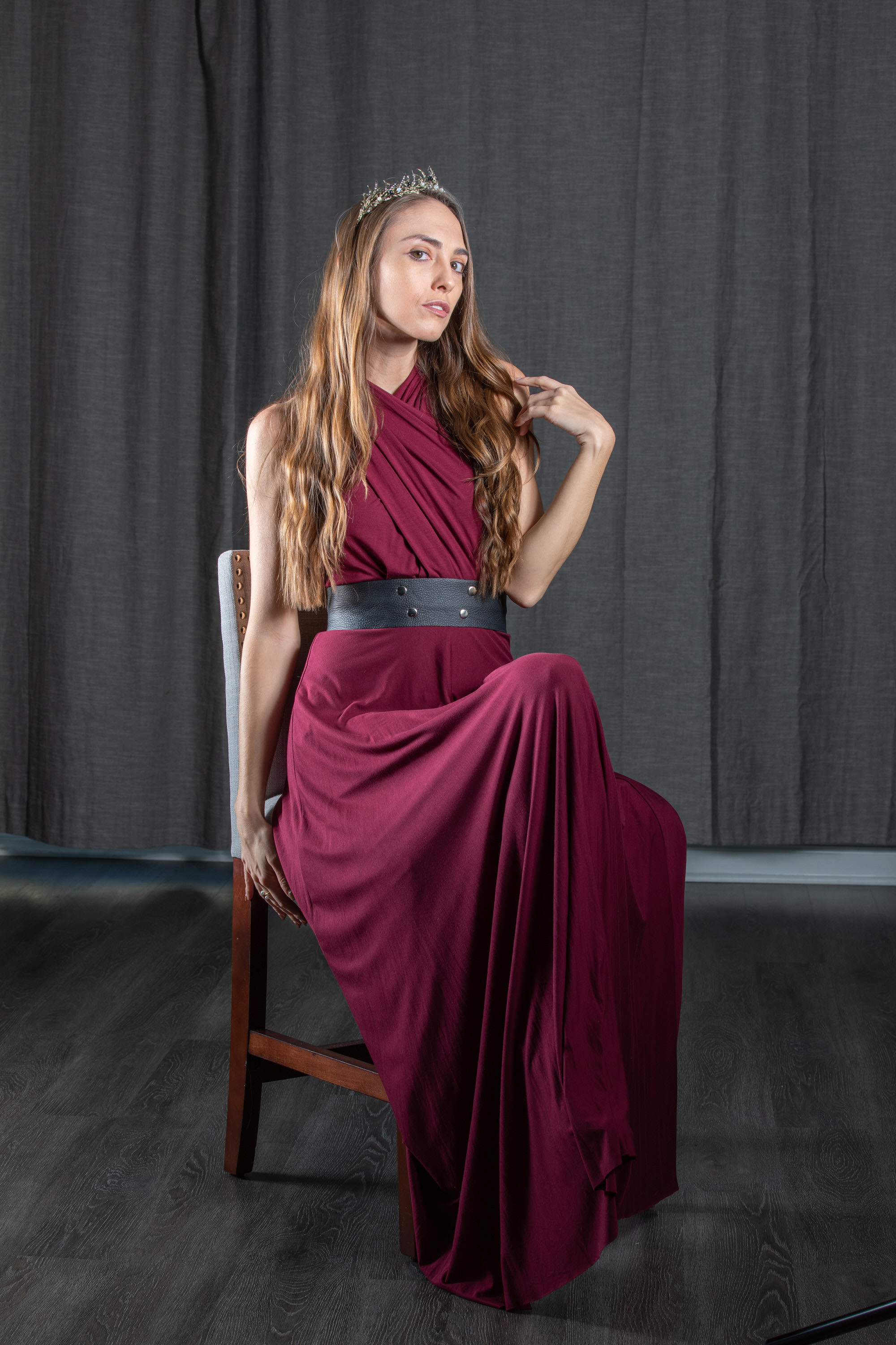 a woman in a long, flowing, maroon dress sits on a wooden chair. she has long brown hair, a crown on her head, and a black belt around her waist. she is posing for the camera against a dark grey backdrop. the lighting is soft and diffused, creating a flattering and elegant mood.